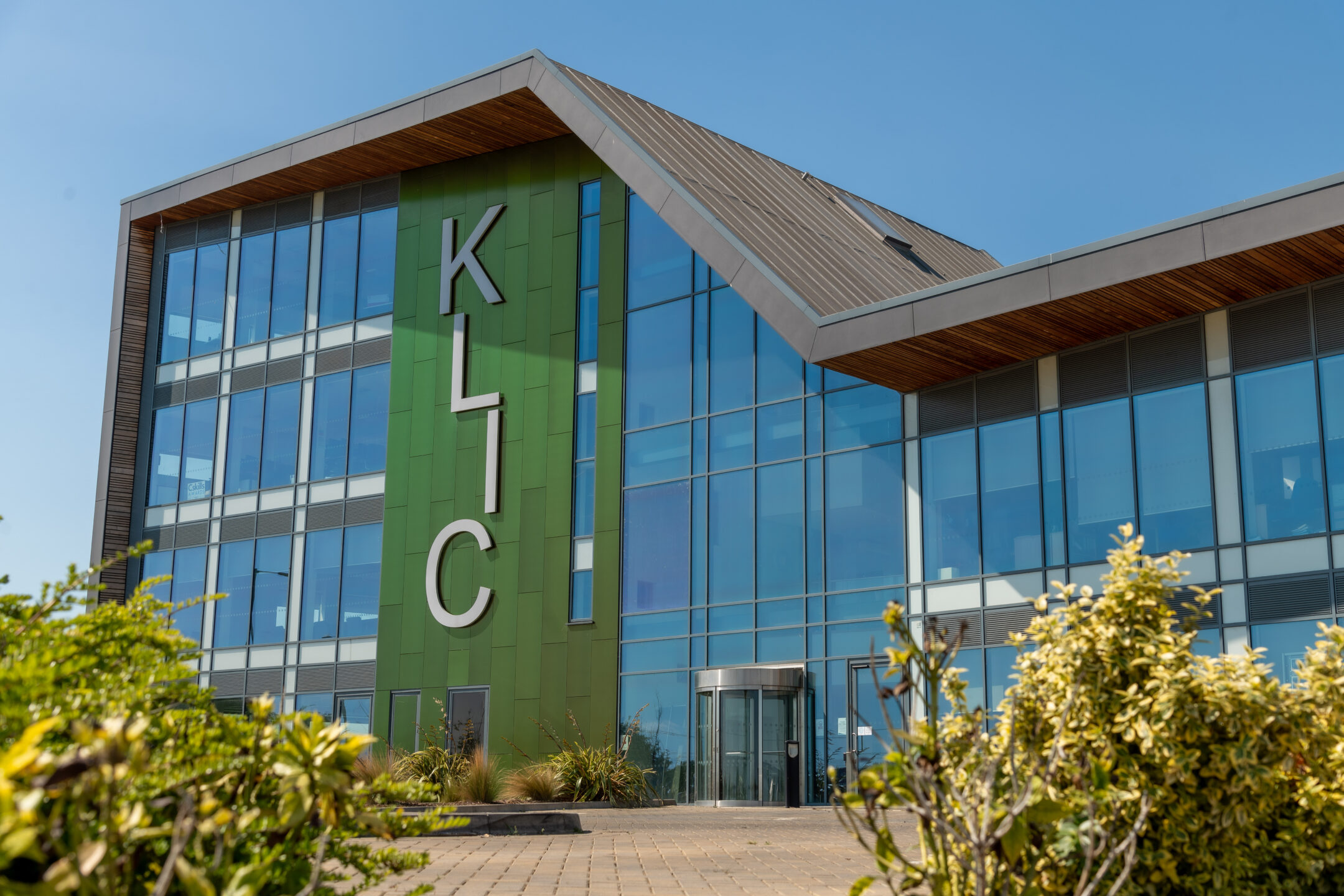 An outside view of the King's Lynn Innovation Centre building (KLIC). There are bushes in front of a 4 storey building with tall windows. The letters "KLIC" run vertically down the front of the building on green metal panelling. This photo is used for the contact page.