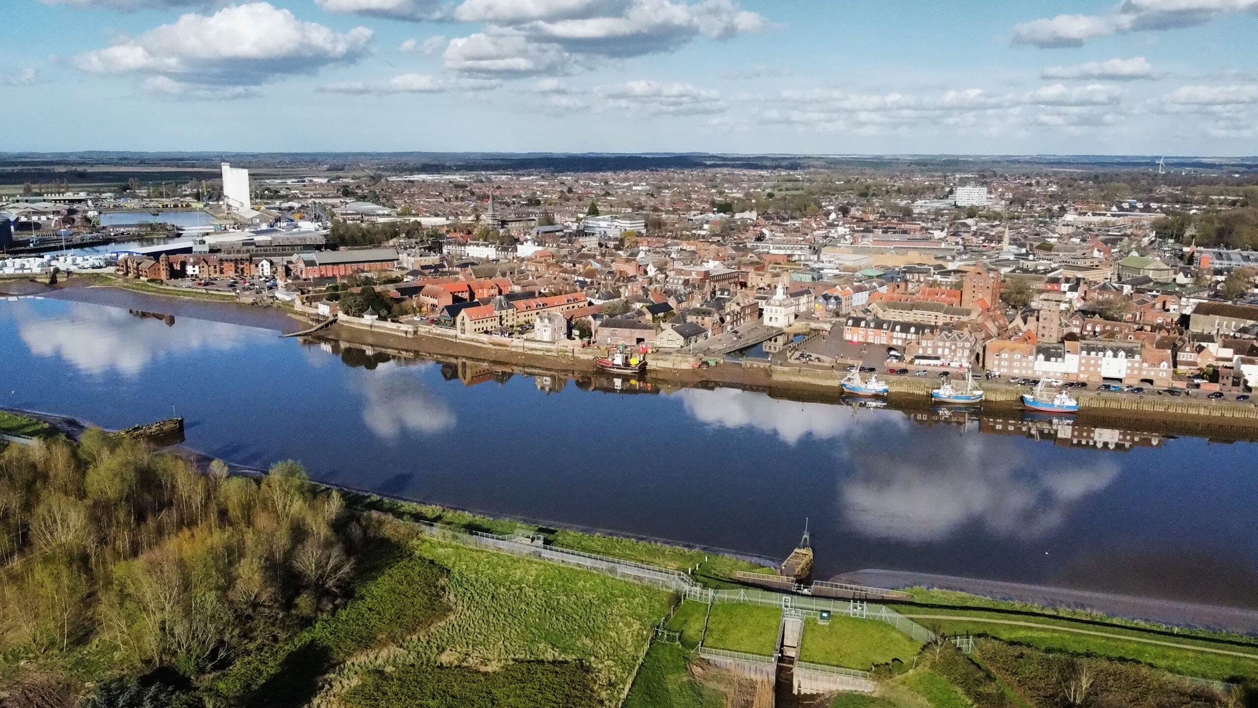 A wide aerial shot of the quay in King's Lynn. A wide river separates greenery and lots of buildings. This is part of the location for the King's Lynn Enterprise Park.